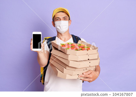 Delivery man employee wearing protective mask holding food order pizza boxes and showing mobile phone with blank screen, isolated on purple background, copy space for advertisement. Delivery man employee wearing protective mask holding food order pizza boxes and showing mobile phone with blank screen, isolated on purple background, copy space for advertisement. 104167807