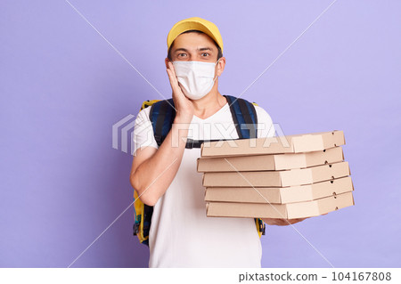 Excited young Caucasian delivery man in protective mask holding pizza boxes isolated on purple background, looking at camera with amazement, keeps hand on cheek, looking at camera. 104167808