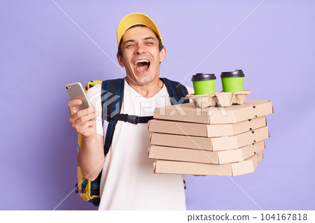 Extremely happy man courier in yellow cap and T-shirt holding food order pizza boxes and coffee to go, standing with phone in hands, screaming happily, isolated on purple background. Extremely happy man courier in yellow cap and T-shirt holding food order pizza boxes and coffee to go, standing with phone in hands, screaming happily, isolated on purple background. 104167818