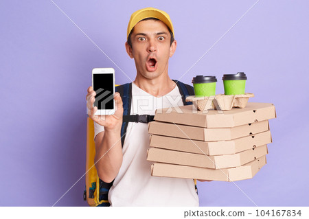 Image of courier holds pizza boxes and takeaway coffee in disposable cups isolated on purple background, showing cell phone with blank black screen, copy space for advertisement. Image of courier holds pizza boxes and takeaway coffee in disposable cups isolated on purple background, showing cell phone with blank black screen, copy space for advertisement. 104167834