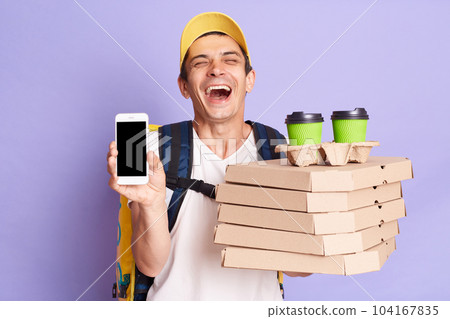 Indoor shot of laughing man in yellow cap and t-shirt, holding pizza and paper cups of coffee, showing mobile phone with empty display, posing isolated on lilac background. 104167835