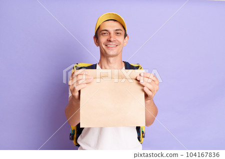 Satisfied delivery man in white t shirt and cap holding paper packet with food isolated over purple background, delivering order, handing packet to client, looking at camera. Satisfied delivery man in white t shirt and cap holding paper packet with food isolated over purple background, delivering order, handing packet to client, looking at camera. 104167836