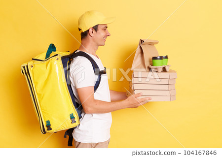 Delivery service. Side view of friendly professional courier white t shirt holding coffee and pizza box, offering drinks and food, looking at client posing isolated on yellow background. Delivery service. Side view of friendly professional courier white t shirt holding coffee and pizza box, offering drinks and food, looking at client posing isolated on yellow background. 104167846