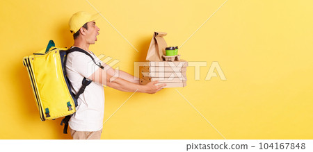 Side view of shocked man in yellow cap and t-shirt, courier holding pizza and paper cups of coffee, handing order to client, posing isolated on yellow background. Side view of shocked man in yellow cap and t-shirt, courier holding pizza and paper cups of coffee, handing order to client, posing isolated on yellow background. 104167848