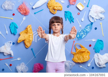 Portrait of serious confident little girl wearing white t shirt posing against blue wall and much plastic garbage around, showing stop gesture. Environmental problems. 104167891
