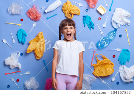 Angry desperate little girl wearing white t shirt posing against blue wall and much plastic garbage around, screaming loud, wants to be heard, shouting about danger for our planet. Angry desperate little girl wearing white t shirt posing against blue wall and much plastic garbage around, screaming loud, wants to be heard, shouting about danger for our planet. 104167892
