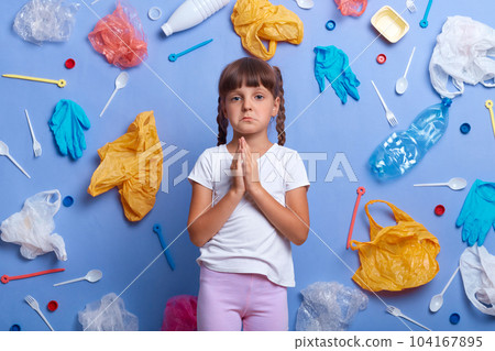 Sad crying little girl dressed casually standing with palms together, praying for Earth, looking at camera with pout lips, prays for ecosystem, poses against blue wall with much waste around. 104167895
