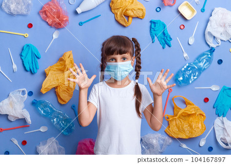 Little girl dressed t shirt and protective mask standing poses against blue wall with much waste around, standing with raised arms, shocked by ecology problem, annoyed by pollution of environment 104167897