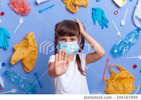 Portrait of little girl with braids wearing white t shirt, keeps hand on head, showing stop gesture with palm, standing against much plastic wastes on blue wall, wants stopping contamination. 104167899