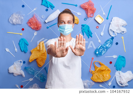 Indoor shot of woman volunteer wearing white t shirt and mask cares about environment, cleans from garbage, concerned with plastic contamination, raises her palms, showing stop gesture. Indoor shot of woman volunteer wearing white t shirt and mask cares about environment, cleans from garbage, concerned with plastic contamination, raises her palms, showing stop gesture. 104167932
