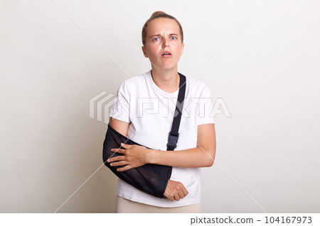 Portrait of Caucasian woman with broken arm, suffers from pain and fracture, wearing arm sling has bad mood, dressed in white t shirt, got into accident, frowning face from terrible pain. Portrait of Caucasian woman with broken arm, suffers from pain and fracture, wearing arm sling has bad mood, dressed in white t shirt, got into accident, frowning face from terrible pain. 104167973