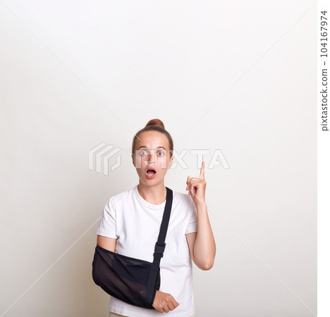 Portrait of shocked astonished woman with bun hairstyle wearing casual t shirt and sling for injured arm posing isolated over white background, pointing up and copy space for advertisement. Portrait of shocked astonished woman with bun hairstyle wearing casual t shirt and sling for injured arm posing isolated over white background, pointing up and copy space for advertisement. 104167974
