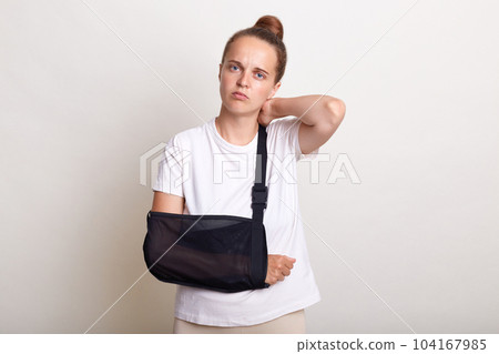 Portrait of sad sick woman with bun hairstyle wearing casual T-shirt standing in arm sling supported on her elbow, feeling pain in neck, looking at camera, suffering in accident, isolated on light Portrait of sad sick woman with bun hairstyle wearing casual T-shirt standing in arm sling supported on her elbow, feeling pain in neck, looking at camera, suffering in accident, isolated on light 104167985