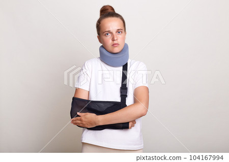 Image of sick injured woman with bun hairstyle wearing casual T-shirt and Shants collar, looking at camera, holding her hands, felling pain, standing in arm sling isolated on light gray background. Image of sick injured woman with bun hairstyle wearing casual T-shirt and Shants collar, looking at camera, holding her hands, felling pain, standing in arm sling isolated on light gray background. 104167994