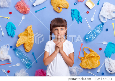 Indoor shot of cute sad little girl wearing white t shirt posing against blue wall and much plastic garbage around, keeps hands together, worries about our planet pollution. 104168024