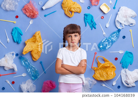 Almost crying little girl with pout lips wearing white t shirt posing against blue wall and much plastic garbage around, keeps hands crossed, looking at camera, worries of danger for our planet. Almost crying little girl with pout lips wearing white t shirt posing against blue wall and much plastic garbage around, keeps hands crossed, looking at camera, worries of danger for our planet. 104168039