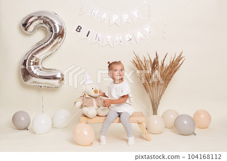 Portrait of little cute blond girl sitting on wooden bench among balloons, celebrating two years, posing in festive room with decoration and big number two made of inflatable foil balloon. 104168112