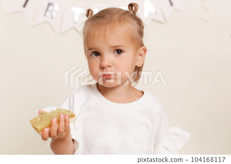 Pretty little infant girl with bun hair buns dressed white attire enjoys delicious cake for her birthday, posing against white wall, looking at camera. Children's holiday. Happy childhood. Pretty little infant girl with bun hair buns dressed white attire enjoys delicious cake for her birthday, posing against white wall, looking at camera. Children's holiday. Happy childhood. 104168117