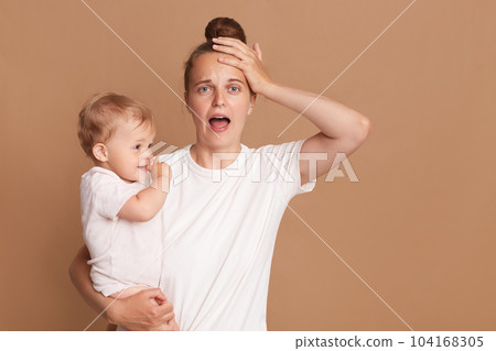 Indoor shot of young tired shocked mother holding a child in her arms, female wearing white casual style T-shirt posing with a baby isolated on brown background, screaming, keeps hand on head. 104168305