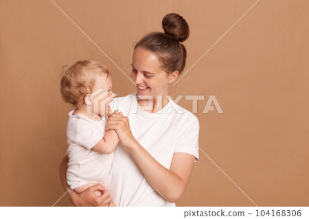 Indoor shot of mother playing with baby girl, happy family having fun at home, cheerful sweet kid in mom's arms, mom and child isolated over brown background. Indoor shot of mother playing with baby girl, happy family having fun at home, cheerful sweet kid in mom's arms, mom and child isolated over brown background. 104168306
