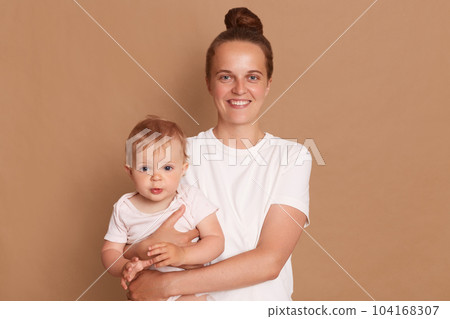 Portrait of satisfied smiling mother holding infant baby girl, happy family having fun together, adorable kid in mom's arms, mom and child isolated over brown background. Portrait of satisfied smiling mother holding infant baby girl, happy family having fun together, adorable kid in mom's arms, mom and child isolated over brown background. 104168307
