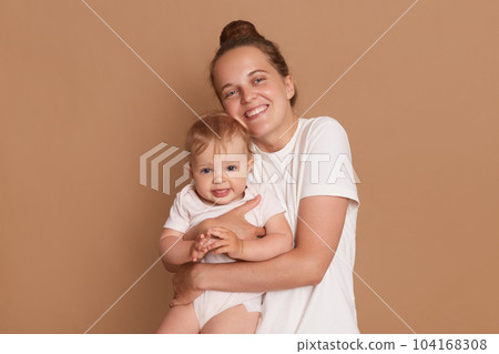 Horizontal shot of smiling satrisfied woman wearing white casual style T-shirt embracing little baby girl, looking at camera with happy expression, posing isolated over brown background. Horizontal shot of smiling satrisfied woman wearing white casual style T-shirt embracing little baby girl, looking at camera with happy expression, posing isolated over brown background. 104168308