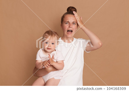 Indoor shot of stressed depressed young mother with bun hairstyle holding her toddler daughter, screaming, being tired and exhausted, touching head, needs rest, standing isolated over brown background Indoor shot of stressed depressed young mother with bun hairstyle holding her toddler daughter, screaming, being tired and exhausted, touching head, needs rest, standing isolated over brown background 104168309