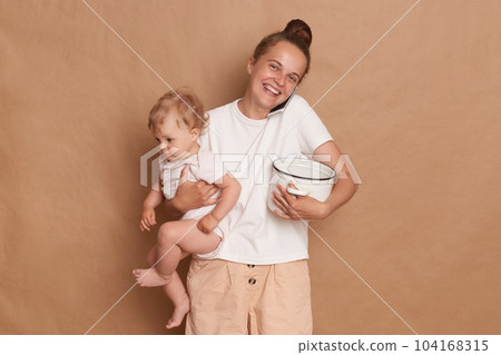 Indoor shot of cheerful joyful Caucasian mother with bun hairstyle holding her infant daughter and pot with porridge or soup, talking phone isolated over brown background. Indoor shot of cheerful joyful Caucasian mother with bun hairstyle holding her infant daughter and pot with porridge or soup, talking phone isolated over brown background. 104168315