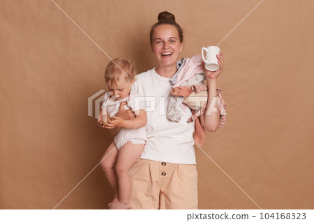 Indoor shot of smiling happy woman mother wearing white T- shirt standing with her little daughter in hands isolated over brown background, drinking coffee in the morning. 104168323