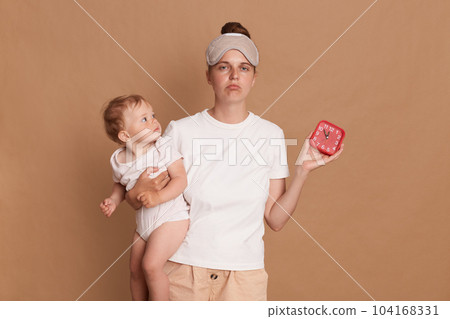 Indoor shot of sad upset Caucasian woman with dark hair wearing sleeping standing with her baby daughter in hands and showing alarm clock, sleepy mother needs more rest. 104168331