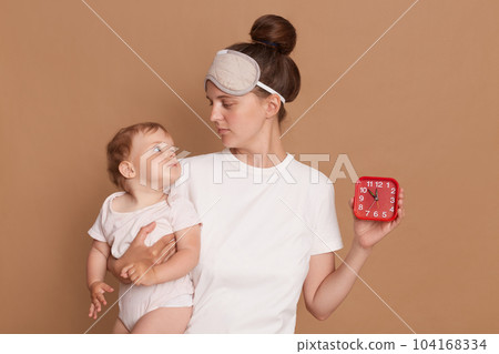 Indoor shot of dark haired mother with sleeping mask holding baby daughter and red alarm clock isolated over brown background, looking at kid, time to wake up and doing routine procedures. 104168334