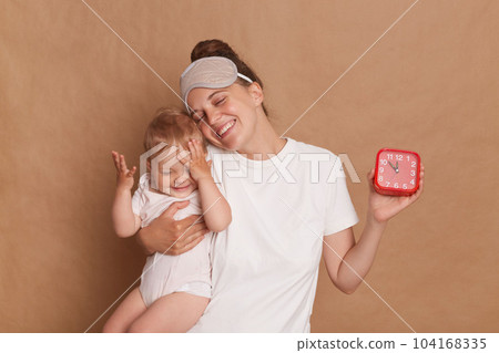 Portrait of satisfied delighted woman wearing white T- shirt standing with her little daughter in hands isolated over brown background, showing red alarm clock, family waking up in good mood. 104168335