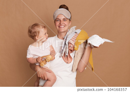 Smiling young mother with bun hairstyle holding her toddler daughter, clothing and diaper, looking at camera with toothy smile, enjoying her maternity leave, posing isolated over brown background. Smiling young mother with bun hairstyle holding her toddler daughter, clothing and diaper, looking at camera with toothy smile, enjoying her maternity leave, posing isolated over brown background. 104168357