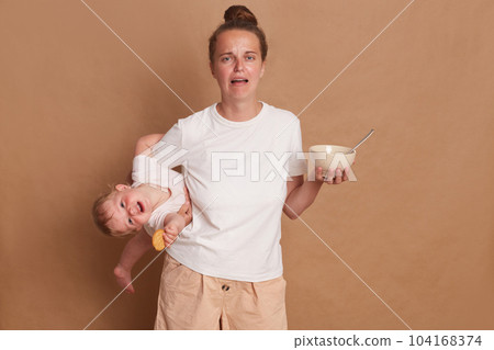 Portrait of shocked crying screaming woman wearing white T- shirt standing with her little daughter in hands isolated over brown background, trying to feed her child. Portrait of shocked crying screaming woman wearing white T- shirt standing with her little daughter in hands isolated over brown background, trying to feed her child. 104168374