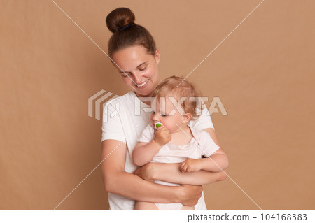 Portrait of smiling loving gentle mother hugging baby daughter isolated over brown background, mommy looking at kid with smile, female wearing white shirt. 104168383