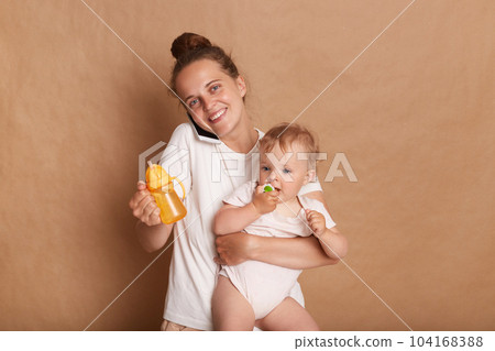 Portrait of attractive happy woman wearing casual clothing standing with her baby child isolated over brown background, looking at camera and talking smart phone. Portrait of attractive happy woman wearing casual clothing standing with her baby child isolated over brown background, looking at camera and talking smart phone. 104168388