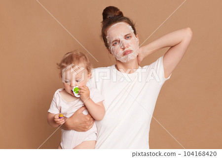 Indoor shot of sad unhealthy woman wearing white t shirt doing cosmetic procedures, feels unwell, has pain neck, looking at camera with sad expression, holding baby daughter in hands. Indoor shot of sad unhealthy woman wearing white t shirt doing cosmetic procedures, feels unwell, has pain neck, looking at camera with sad expression, holding baby daughter in hands. 104168420
