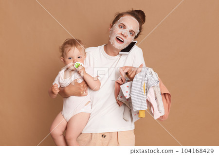 Amazed woman wearing white t shirt doing cosmetic procedures, holding baby daughter in hands and kid's clothing, talking via cell phone with her friend, isolated over brown background. 104168429