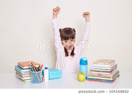 Extremely happy excited little girl with braids sitting at desk posing isolated over white background, being among school supplies, being glad, raised arms, break starting, the lesson is over. Extremely happy excited little girl with braids sitting at desk posing isolated over white background, being among school supplies, being glad, raised arms, break starting, the lesson is over. 104168692