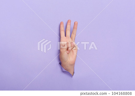 Indoor shot of human's hand sticks out of hole in paper isolated on purple background. Three finger sign, copy space for advertisement. 104168808
