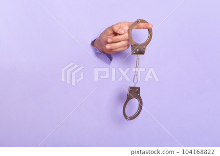 Person's hand holding handcuffs in purple paper hole, silver shackle in human's hand breaking through lilac background. Criminality, misdemeanor. Person's hand holding handcuffs in purple paper hole, silver shackle in human's hand breaking through lilac background. Criminality, misdemeanor. 104168822
