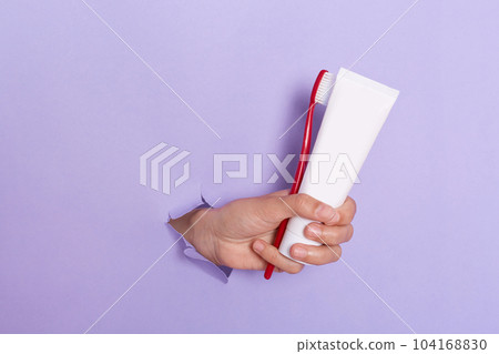 Indoor shot of person's holding toothbrush and toothpaste isolated on purple background, human hand breaking through lilac paper torn, oral protection. 104168830