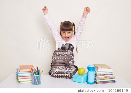 Extremely happy positive little girl with braids standing near desk with raised arms, screaming happily, being glad that her lessons are over, posing isolated over white background. Extremely happy positive little girl with braids standing near desk with raised arms, screaming happily, being glad that her lessons are over, posing isolated over white background. 104169149