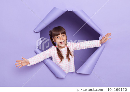 Indoor shot of excited happy little girl with pigtails wearing striped shirt breaks though paper hole with raised arms, imagines she is a plane, screaming happily. 104169150