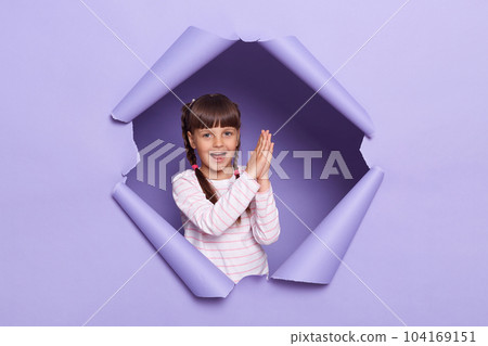 Indoor short of dark haired little girl with pigtails wearing striped shirt breaks through purple paper background, looking at camera and clapping hands, expressing excitement. Indoor short of dark haired little girl with pigtails wearing striped shirt breaks through purple paper background, looking at camera and clapping hands, expressing excitement. 104169151
