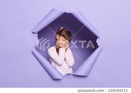 Portrait of sleepy tired adorable little girl with braids looking through breakthrough of purple background, standing with closed eyes, leaning on her palms, feels sleepless. Portrait of sleepy tired adorable little girl with braids looking through breakthrough of purple background, standing with closed eyes, leaning on her palms, feels sleepless. 104169152