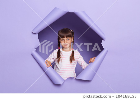 Portrait of positive happy little girl wearing striped shirt posing in torn purple paper hole, expressing optimism, looking at camera with calm expression. Portrait of positive happy little girl wearing striped shirt posing in torn purple paper hole, expressing optimism, looking at camera with calm expression. 104169153