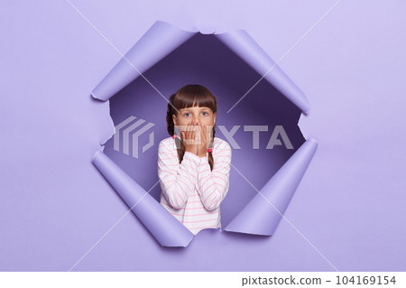 Horizontal shot of shocked scared little girl with braids looking through breakthrough of purple background, sees something astonished, covering mouth with palms, looking with big eyes. Horizontal shot of shocked scared little girl with braids looking through breakthrough of purple background, sees something astonished, covering mouth with palms, looking with big eyes. 104169154
