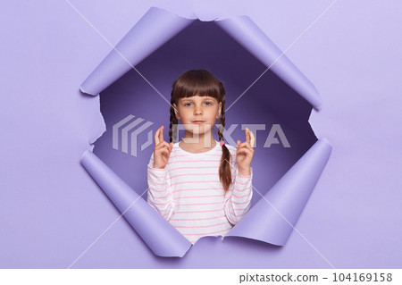 Indoor shot of calm charming adorable little girl with braids looking through breakthrough of purple background, standing with crossed fingers, looks hopeful. Indoor shot of calm charming adorable little girl with braids looking through breakthrough of purple background, standing with crossed fingers, looks hopeful. 104169158