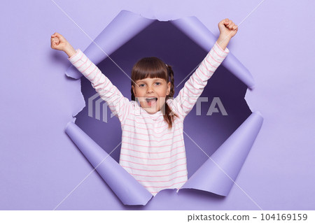 Portrait of amazed excited little girl with braids wearing striped shirt posing in torn paper wall, kid standing with raised arms, screaming happily, celebrating her success. 104169159